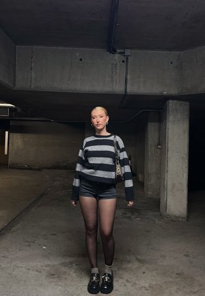 Suéter a rayas gris y negro, pantalones cortos negros, pantimedias transparentes, zapatos negros. Los accesorios incluyen un bolso de leopardos. Fondo de concreto.