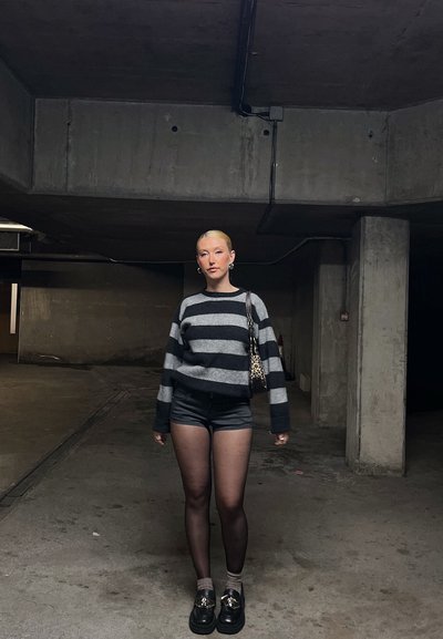 Suéter a rayas gris y negro, pantalones cortos negros, pantimedias transparentes, zapatos negros. Los accesorios incluyen un bolso de leopardos. Fondo de concreto.