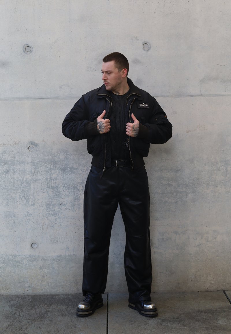 Veste bomber noire avec fermeture éclair et détail logo par-dessus un pull en tricot foncé. Pantalon en cuir noir et chaussures plateformes noires brillantes.