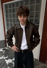 Veste bomber en cuir marron avec col à revers, fermeture éclair, poignets côtelés et poches latérales, portée sur un polo blanc et un pantalon foncé.