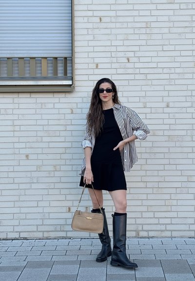 Mujer con cabello largo, vestida con vestido negro, camisa de rayas, botas negras, gafas de sol, sosteniendo un bolso beige, de pie junto a una pared de ladrillos blanca.