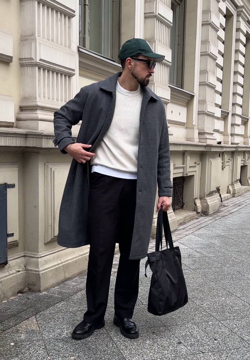 Homme portant un pardessus gris, un pull blanc, un pantalon noir, une casquette verte, des lunettes de soleil, tenant un sac fourre-tout noir, debout dans une rue de la ville devant une façade de bâtiment ornée.