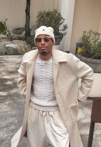 Manteau en laine beige porté sur un pull blanc à torsades, associé à un pantalon de survêtement crème. Les accessoires comprennent un bonnet blanc et des lunettes de soleil noires.