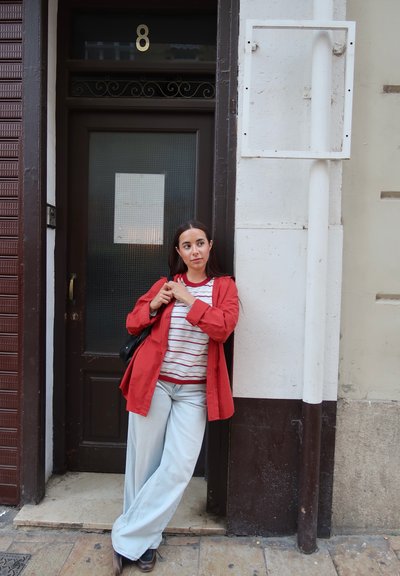 Mujer con chaqueta roja y camisa a rayas apoyada en una puerta negra con el número 8 en una acera de la ciudad.