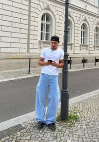 Lekka biała koszulka, oversize'owe jasnoniebieskie dżinsy i czarne buty. Osoba trzyma telefon, opierając się o latarnię.