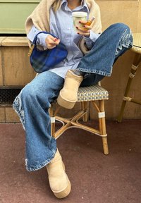 Μπεζ σουέτ πλατφόρμα μποτάκια, μπλε καρό oversized πουκάμισο, φθαρμένα μπλε τζιν, κρατώντας ένα πράσινο χάρτινο ποτήρι και μια μπλε τσάντα με μοτίβο, καθισμένος σε μια καρέκλα.