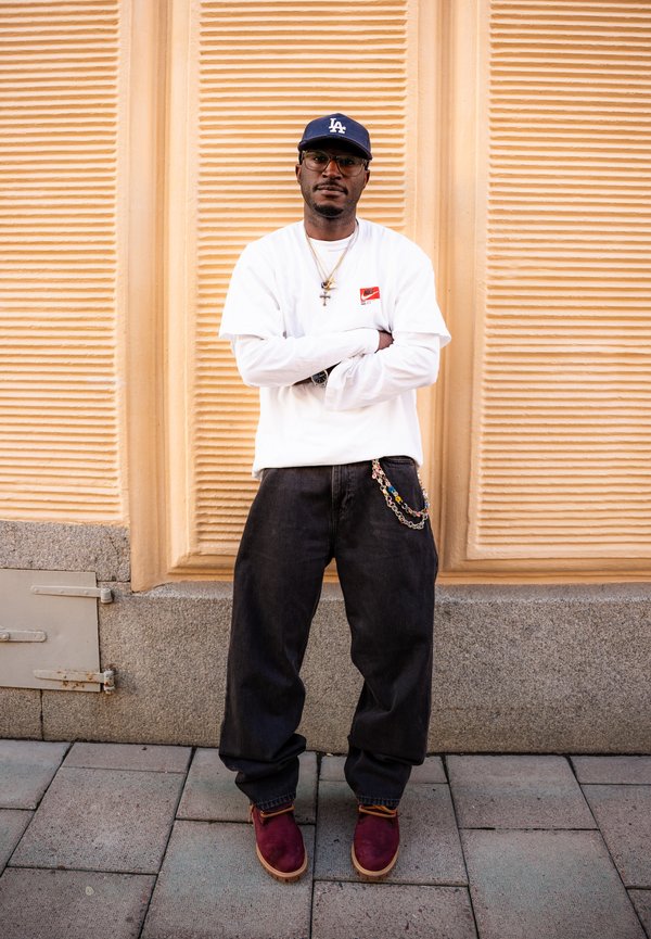 Homme portant un t-shirt Nike blanc à manches longues, un pantalon noir ample avec une chaîne, des chaussures bordeaux, des lunettes et une casquette de baseball bleue, debout les bras croisés.