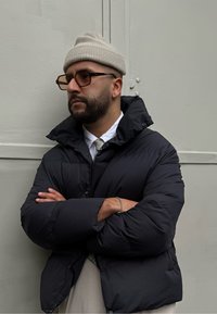 Μαύρο μπουφάν με ψηλό γιακά, φορεμένο πάνω από λευκό πουκάμισο και γραβάτα. Τα αξεσουάρ περιλαμβάνουν μια μπεζ ζακέτα και καφέ γυαλιά. Ανοιχτό γκρίζο φόντο.