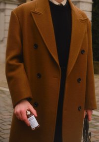 Manteau en laine marron à double boutonnage avec des boutons noirs, porté sur un pull noir. Main tenant une bouteille en verre et un petit sac à main noir.