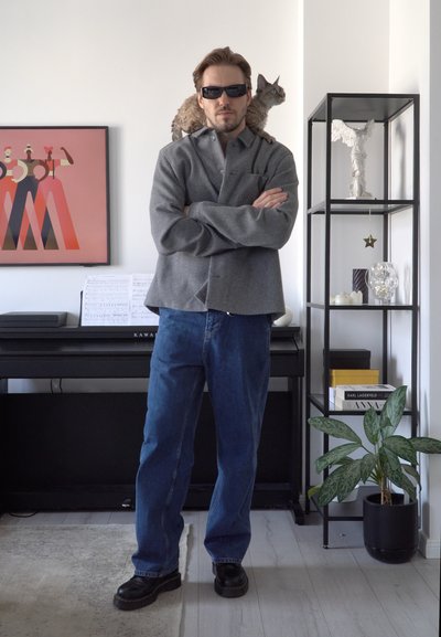 Camisa de botones gris, jeans azules y zapatos negros. Entorno interior con un gato en los hombros, un piano y una estantería decorativa al fondo.