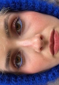 Gros plan sur un visage à la peau claire, portant un eye-liner bleu et un rouge à lèvres mat rose. Encadré par un bonnet tricoté bleu, avec des cheveux courts et clairs.