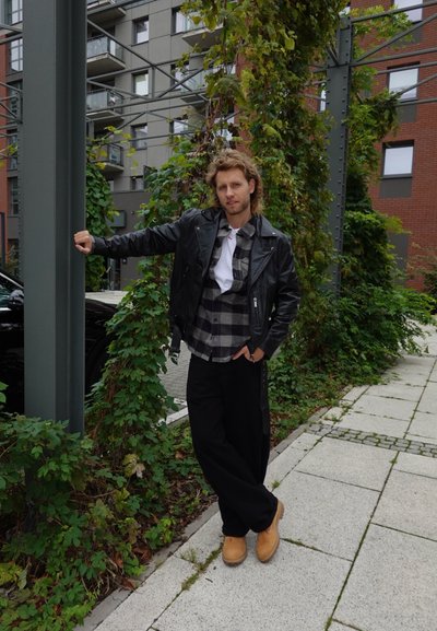 Chaqueta de cuero negra sobre una camisa de cuadros gris y negra, combinada con pantalones holgados negros y zuecos color marrón claro; follaje verde y edificios grises en el fondo.