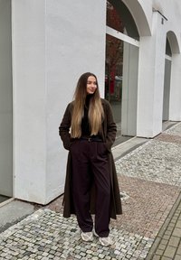 Femme aux cheveux longs et raides portant un long manteau marron, un haut noir, un pantalon large noir et des baskets blanches, debout sur un trottoir pavé.