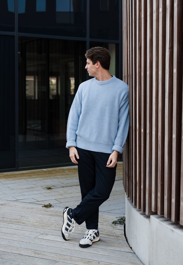 Suéter de punto azul claro con textura acanalada, combinado con pantalones oscuros y zapatillas en blanco y negro. De pie contra una pared de madera.