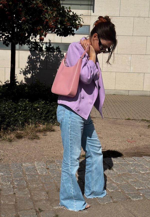Femme portant des lunettes de soleil, une veste violette et un jean évasé bleu clair, portant un sac à main rose à l'épaule, debout à l'extérieur près d’un mur.