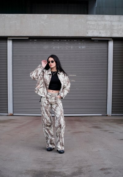Mujer con chaqueta y pantalones estampados, top corto negro y gafas de sol, de pie frente a unas puertas correderas metálicas cerradas de color gris.