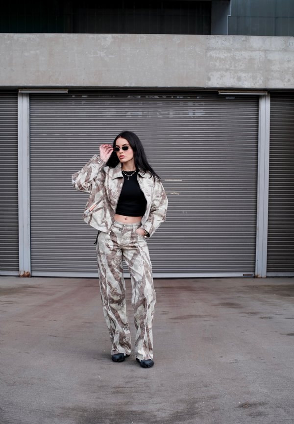 Kvinne iført mønstret jakke og bukser med svart crop-top og solbriller, står foran lukkede grå metallrulleskodder.