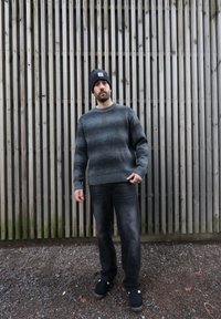 Pull gris en tricot avec une texture horizontale, un col rond et des poignets côtelés. Associé à un jean foncé et un bonnet bleu. Debout devant des lattes de bois.