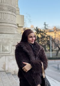 Ψεύτικο καφέ παλτό με φαρδύ γιακά, φορεμένο πάνω από ένα σκούρο ζιβάγκο. Μια χρυσή τσάντα ώμου φαίνεται. Λείες, κυματιστές σκούρες καφέ τρίχες, φωτεινό φόντο.