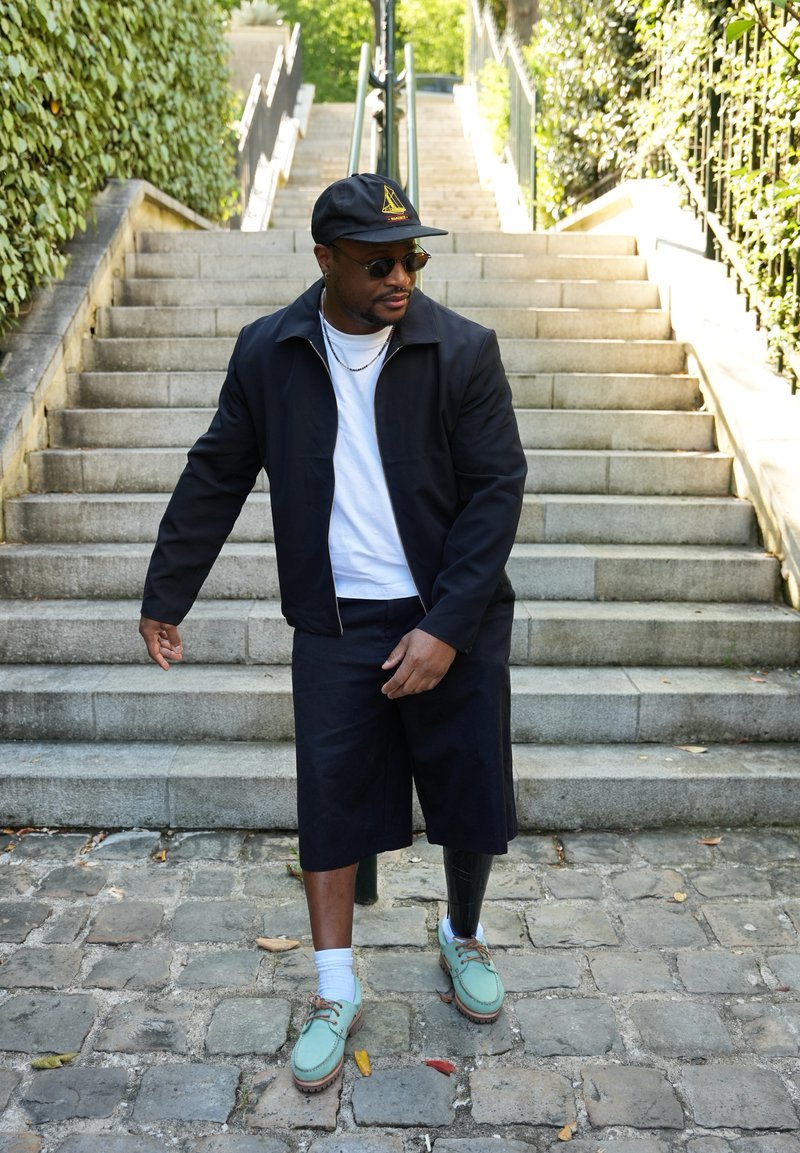 Homme avec jambe prothétique portant une casquette noire, des lunettes de soleil, une veste noire, une chemise blanche et des chaussures turquoise, debout sur des marches en pavés en extérieur.