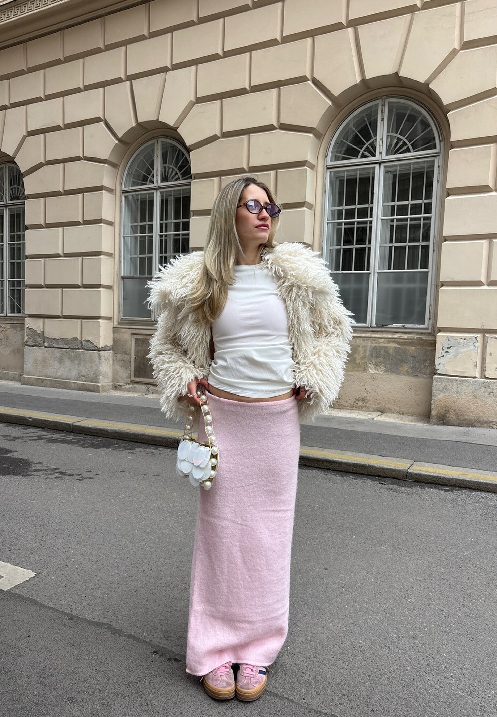 Casaco de pelúcia branca, top branco ajustado, saia longa de malha rosa, mala transparente com detalhes em pérola e sapatos rosas. O fundo apresenta paredes de pedra.
