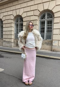 Casaco de pelúcia branca, top branco ajustado, saia longa de malha rosa, mala transparente com detalhes em pérola e sapatos rosas. O fundo apresenta paredes de pedra.