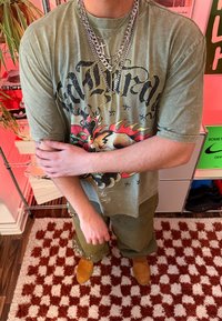 Personne portant un t-shirt graphique vert olive, des chaînes argentées superposées, un pantalon vert olive et des bottes beige, debout sur un tapis à carreaux rouges et blancs à l'intérieur.