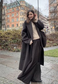 Długi czarny wełniany płaszcz na beżowym swetrze, szerokie czarne spodnie oraz pikowana czarna torba. Gładka faktura i minimalistyczny design.