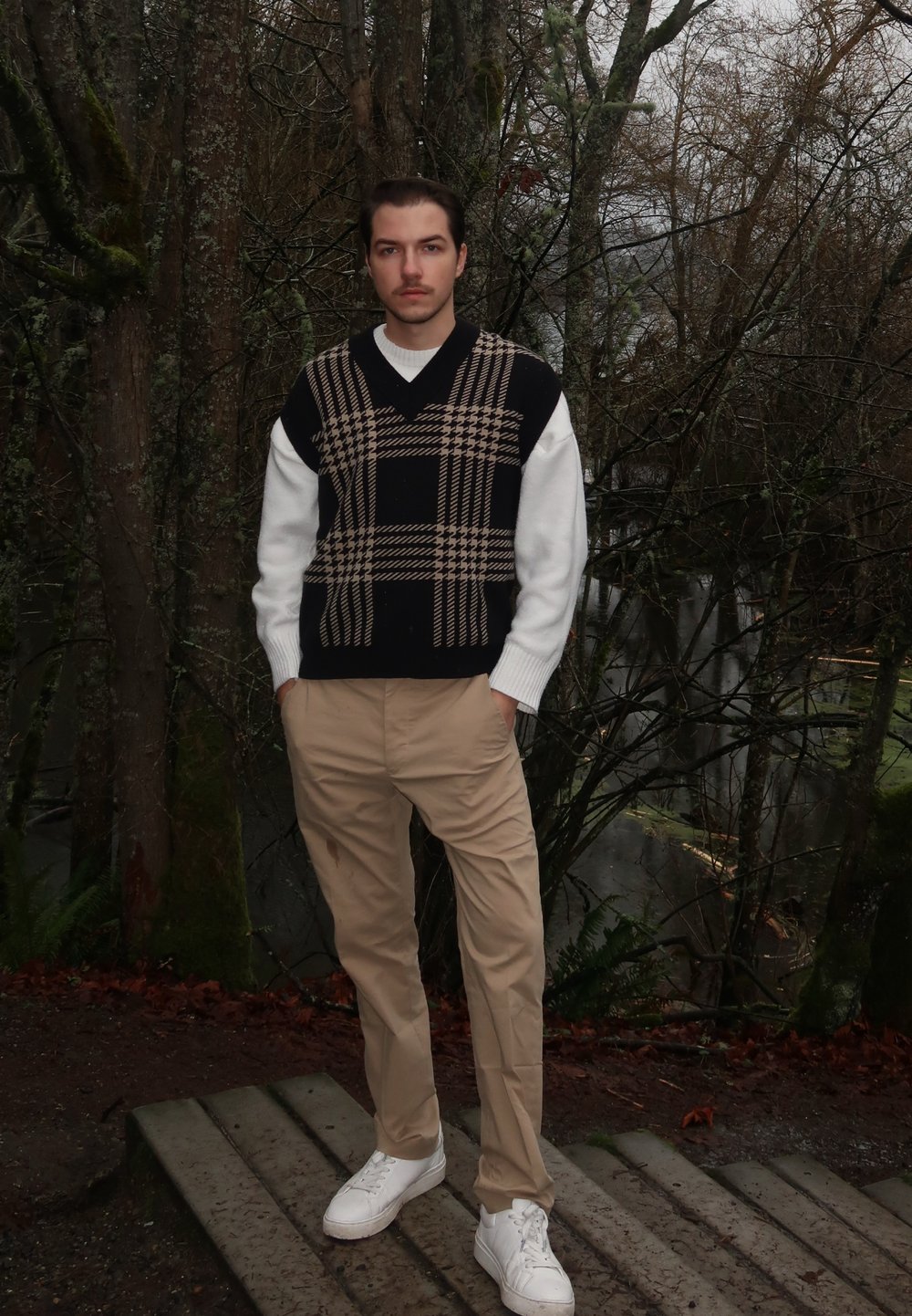 Gilet en maille à motif à carreaux noir et beige porté sur une chemise blanche à manches longues ; associé à un pantalon beige et des baskets blanches.