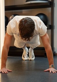 Hvitt treningsantrekk, utfører en armheving på et mørkt gulv. Merkbare detaljer inkluderer en klokke på venstre håndledd og svarte treningsballer i bakgrunnen.