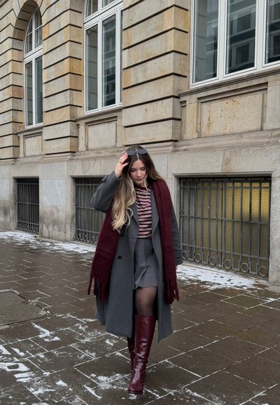 Mujer joven caminando por una acera nevada, vistiendo un abrigo gris, bufanda burdeos, camisa a rayas, falda gris y botas altas burdeos.