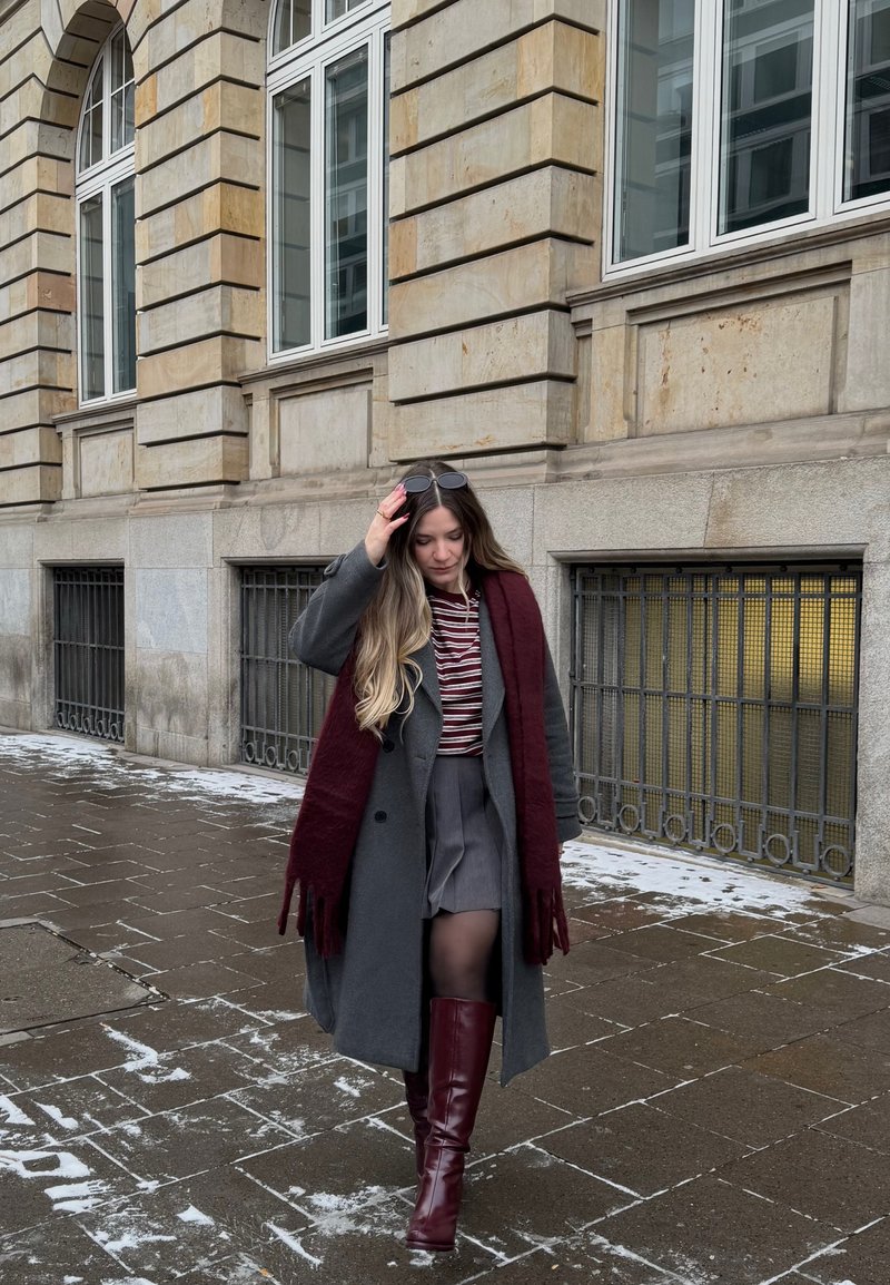 Jeune femme marchant sur un trottoir enneigé, portant un manteau gris, une écharpe bordeaux, une chemise rayée, une jupe grise et de hautes bottes bordeaux.