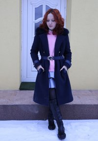 Femme aux cheveux roux portant un manteau noir, un pull rose, une jupe rayée et des bottes noires, debout dans la neige devant un bâtiment jaune avec une porte blanche.
