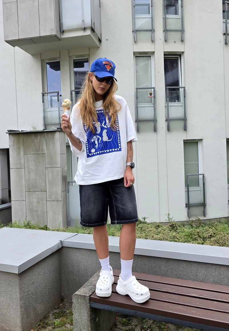 T-shirt oversize blanche avec un graphisme bleu, shorts en denim, sabots blancs et chaussettes blanches, tenant un cornet de glace dans un cadre urbain.