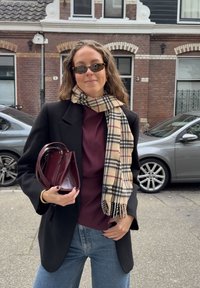 Blazer nero su una maglia bordeaux, abbinato a una sciarpa a quadri beige e nera. Tiene una piccola borsa bordeaux. Ambiente naturale.