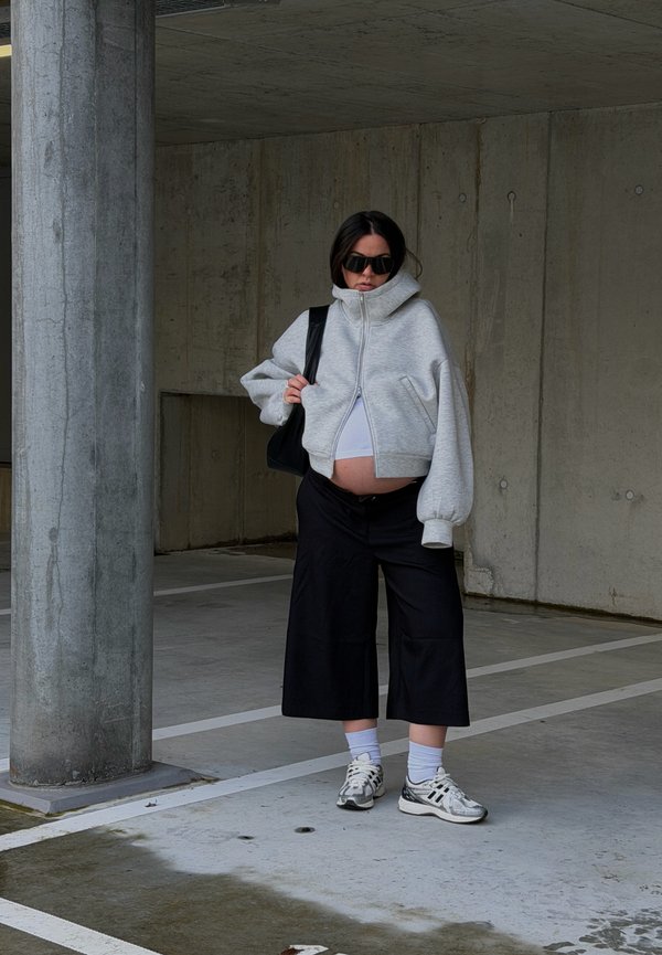 Femme enceinte en sweat à capuche gris zippé, pantalon large noir, baskets et lunettes de soleil, debout dans un parking en béton vide avec une colonne à proximité.