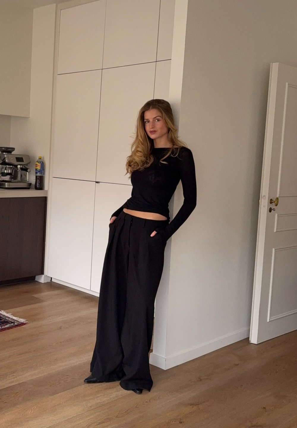 Haut noir à manches longues associé à un pantalon noir ample, debout contre un mur dans un intérieur minimaliste avec un sol en bois.