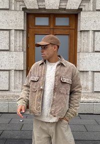 Jeune homme portant une veste en velours côtelé beige et une casquette, debout devant une porte en bois d'un bâtiment en pierre, regardant à gauche avec une main dans la poche.