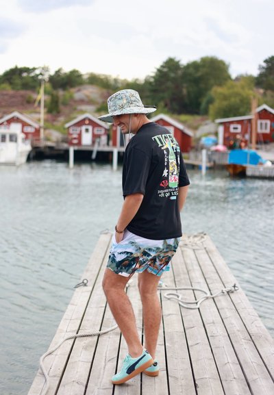 Camiseta gráfica negra con estampado colorido, sombrero de cubo azul claro con patrón, pantalones cortos de baño blancos con diseño de palmera y zapatillas aqua sobre un muelle de madera.