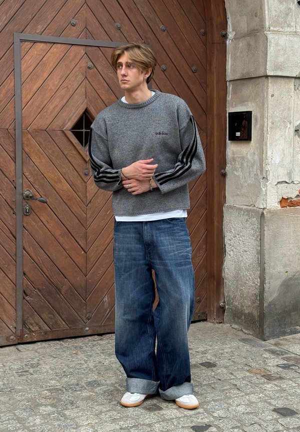 Jeune homme aux cheveux châtain clair portant un sweat gris Adidas, un jean bleu large et des baskets blanches, se tient devant une porte en bois sur un pavé en pierre.