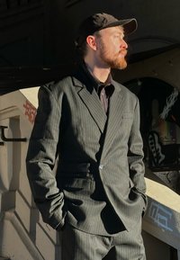 Costume sombre à rayures avec une coupe ajust ée, comprenant un seul bouton et des revers crantés, associé à une chemise boutonnée et une casquette noire.