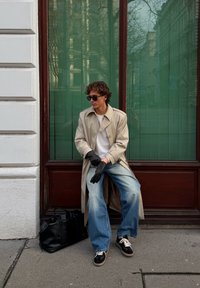 Trench-coat beige, t-shirt blanc, jean bleu à jambes larges, gants noirs et baskets noires. Un sac noir texturé repose au sol.