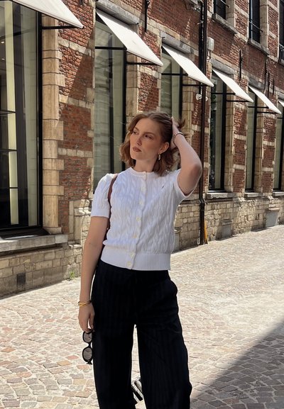 Cárdigan blanco de punto trenzado con mangas cortas y botones amarillos, combinado con pantalones de tiro alto a rayas negras. Textura sólida y ajuste relajado.