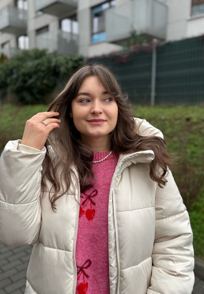 Chaqueta de invierno acolchada de color marfil, suéter rosa con patrón de cerezas y collar de perlas; la modelo se toca el cabello, en un entorno al aire libre con vegetación.