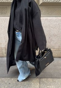 Άτομο που φοράει μαύρο παλτό και πουλόβερ με ευρύχωλο τζιν μπλε παντελόνι, κρατώντας μια μεγάλη μαύρη τσάντα, περπατάει σε πεζοδρόμιο της πόλης.