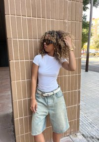 T-shirt branca justa, calções de ganga azul claro com um aspecto desbotado, cinto branco com fivela floral, óculos de sol escuros e cabelo encaracolado.