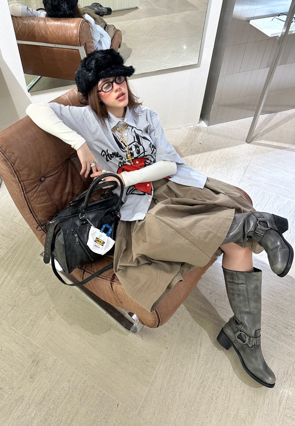 Henkilö istuu ruskeassa nahkatuolissa, päällä harmaa t-paita graafisella printillä, vaaleanbeige hame, harmaat polvisukat ja musta käsilaukku.