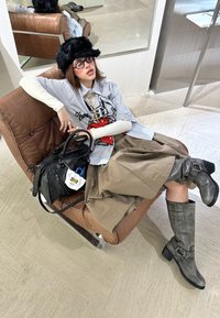 Henkilö istuu ruskeassa nahkatuolissa, päällä harmaa t-paita graafisella printillä, vaaleanbeige hame, harmaat polvisukat ja musta käsilaukku.