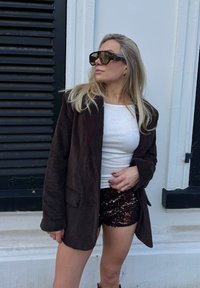 Blazer di velluto a coste marrone, top bianco a coste e pantaloncini neri con paillettes. Occhiali da sole marroni oversize. Sfondo testurizzato con persiane nere.