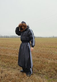 Doudoune noire avec une capuche bordée de fourrure, associée à une jupe noire fluide à rayures blanches, portée par-dessus un pantalon sombre et des bottes.